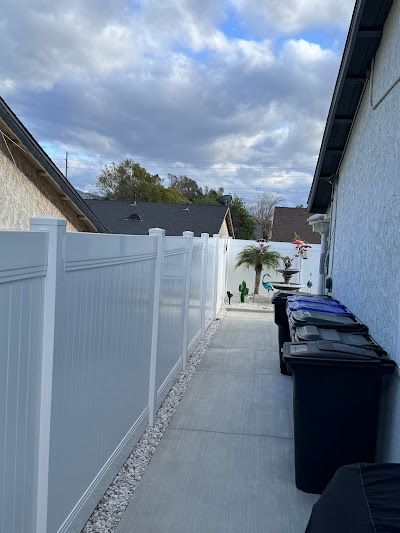 Narrow outdoor side yard with white fence, gray wall, trash bins, and cloudy sky.