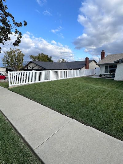 Residential sidewalk beside a green lawn and white fence under a partly cloudy blue sky