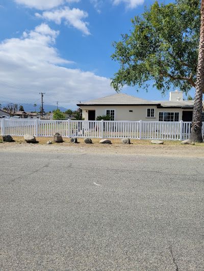 Single-story house behind a white fence with a tree, set beside a quiet road under a cloudy sky