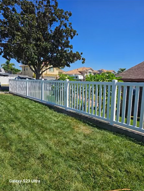 White fence along a green lawn beside a suburban street under a clear blue sky.