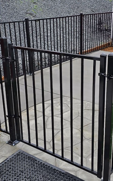 Black metal gate opening onto a concrete ramp beside a gravel area and fenced walkway.