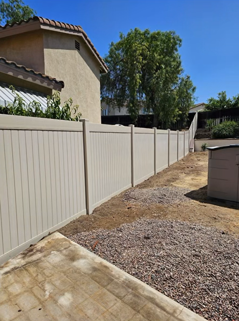 A beige fence along a narrow gravel side yard beside a house, with a tree and trash bin in the distance.