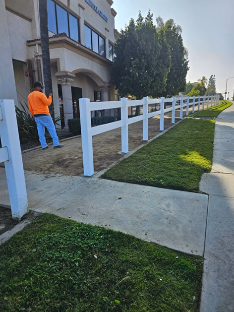 Person in orange jacket on a sidewalk beside a white fence and building under sunny skies