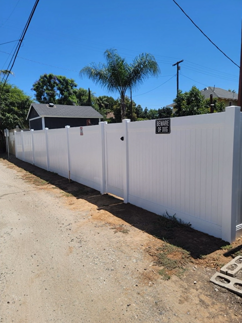 White privacy fence along a dirt roadside under a blue sky, with palm trees and houses behind it.