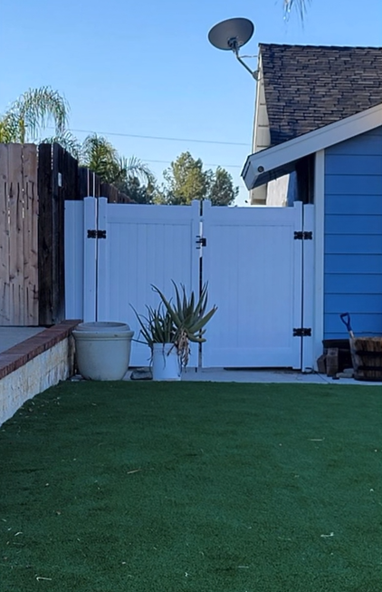 Backyard with white gate, blue house wall, and green artificial grass under a satellite dish.