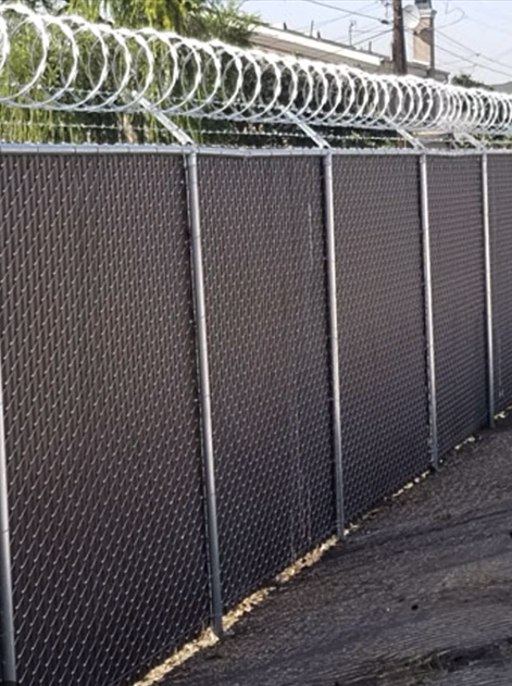 Tall chain-link fence with dark privacy panels topped by razor wire along a paved edge