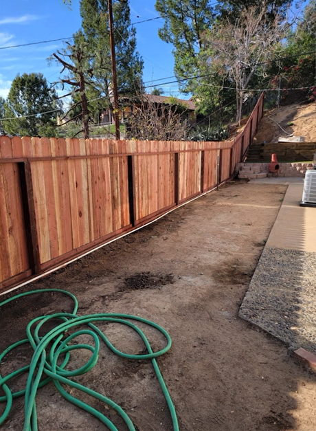 Narrow dirt side yard with a red wooden fence, hose on the ground, and a stone patio edge.