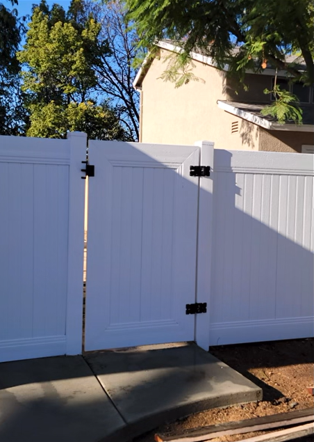 White fence gate in front of a house, with trees and a sidewalk beside it.
