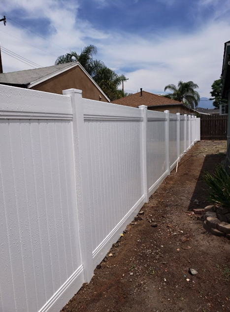 White vinyl fence along a narrow dirt side yard beside suburban houses under a partly cloudy sky