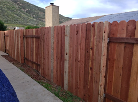 Wooden privacy fence along a sidewalk, with a hillside and house roof in the background.