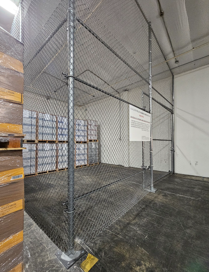 Chain-link fenced warehouse cage with stacked boxes and a posted warning sign under a high ceiling