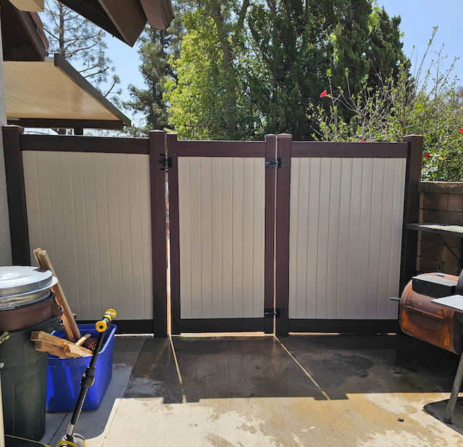 Backyard patio with a beige privacy fence, potted plants, and a blue bin under partial shade
