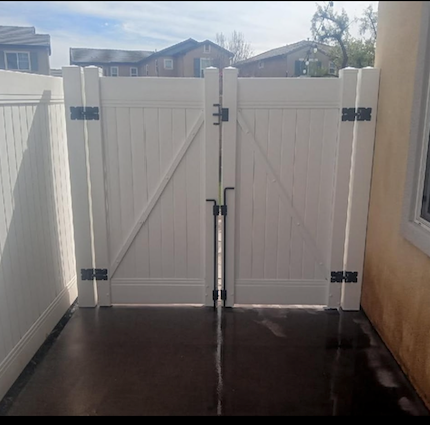 White double gate in a narrow side yard with dark flooring, secured by metal hinges and latch.