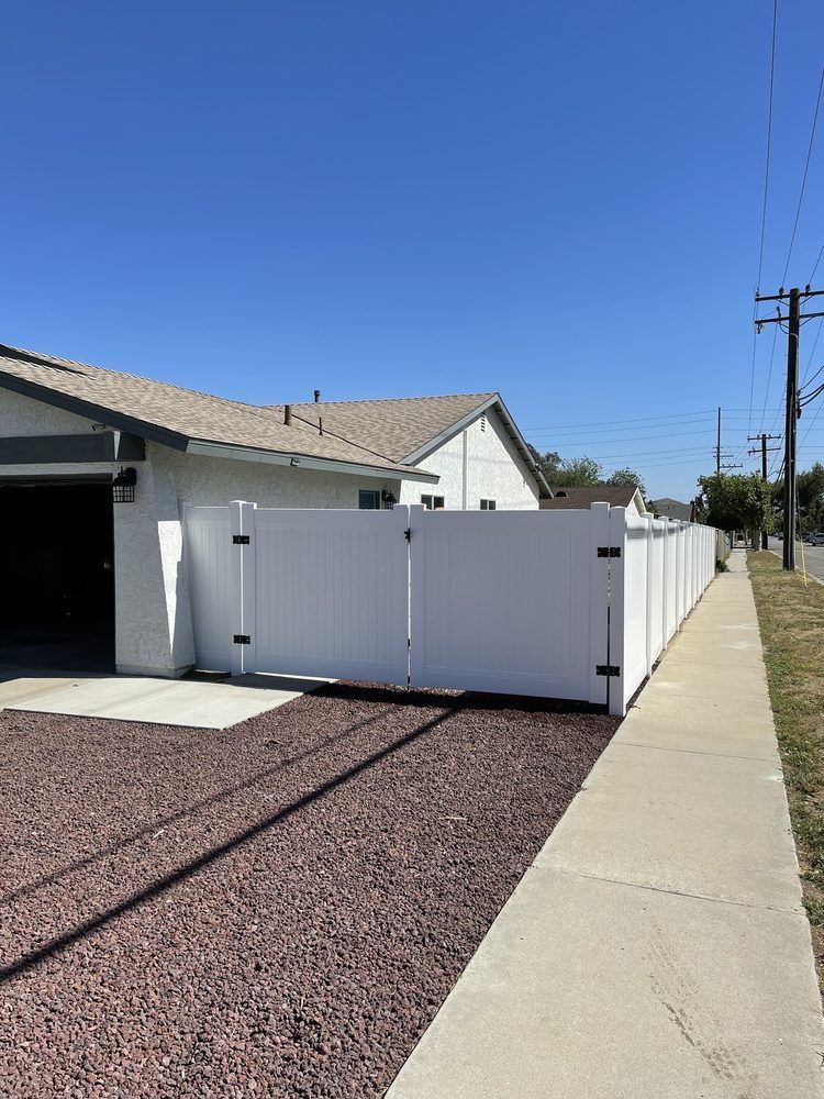 White fence beside a gravel driveway and sidewalk next to a house under a clear blue sky