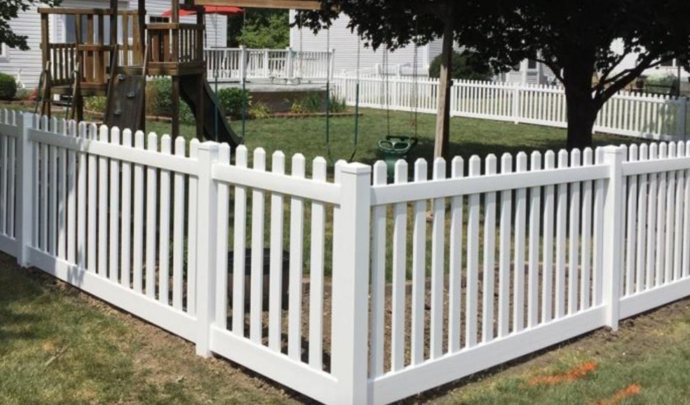 White vinyl fence surrounding a yard with a playground and trees in the background