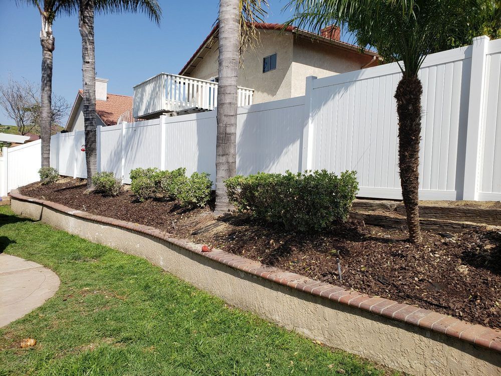 Landscaped yard beside a white fence with palm trees, shrubs, and a concrete curb along a green lawn.