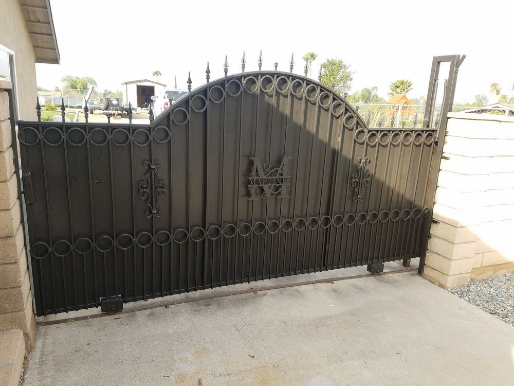Black wrought-iron driveway gate with decorative scrollwork beside a stone wall and paved driveway
