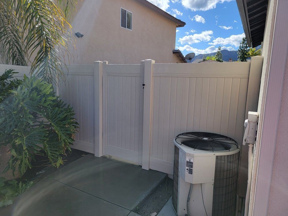 Backyard corner with a white fence, beige house wall, palm plants, and an outdoor AC unit under blue sky.