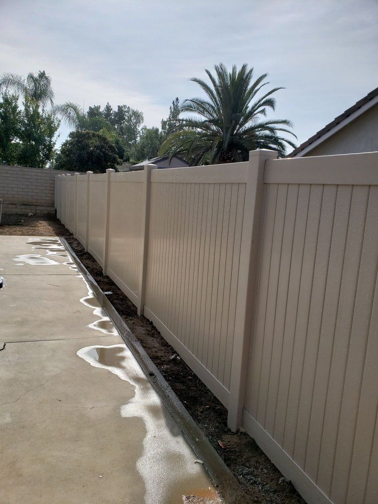 Long beige vinyl fence beside a wet concrete driveway, with trees and a palm visible beyond.