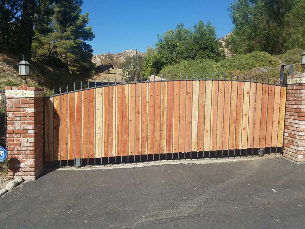 Wooden driveway gate between brick pillars, fronted by asphalt pavement and trees under a clear blue sky