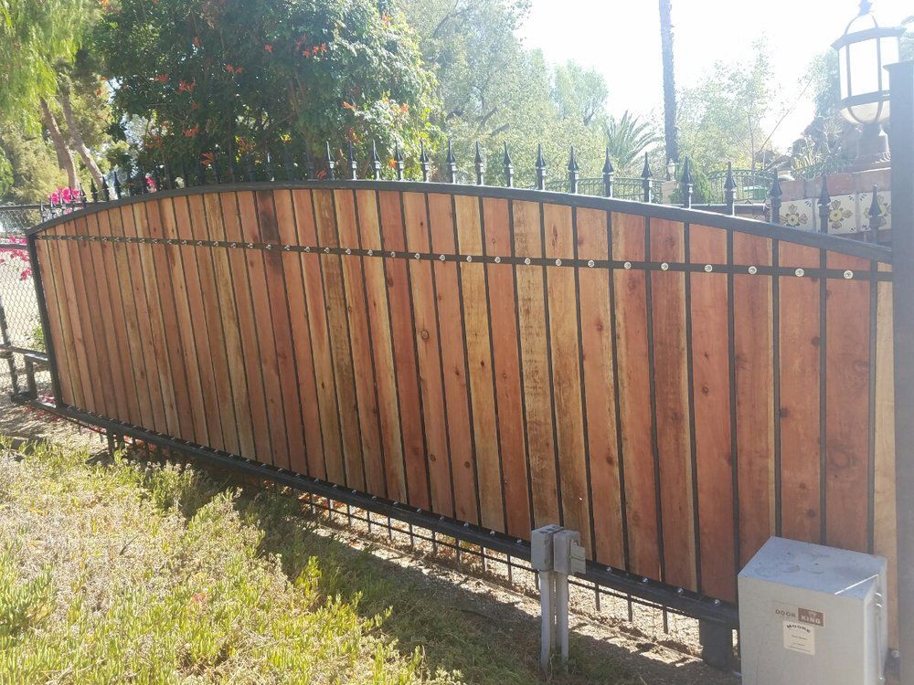 Curved wooden fence with black metal frame in a sunny yard, with grass and trees behind it