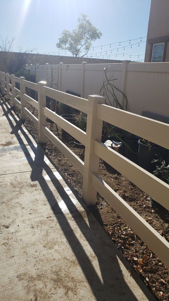 Sunlit beige fence along a gravel path beside a white wall and small tree