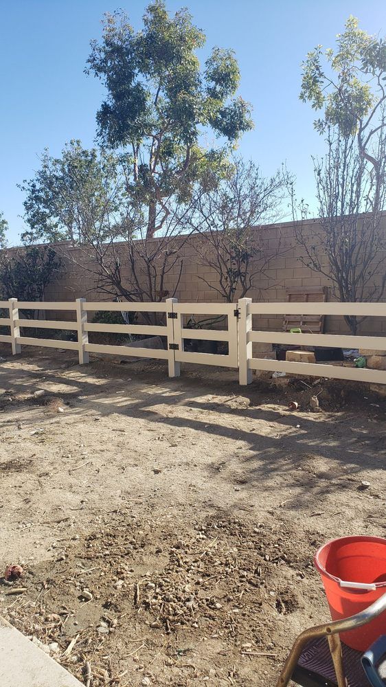 White wooden fence in a dry yard with trees and a red bucket in the foreground.
