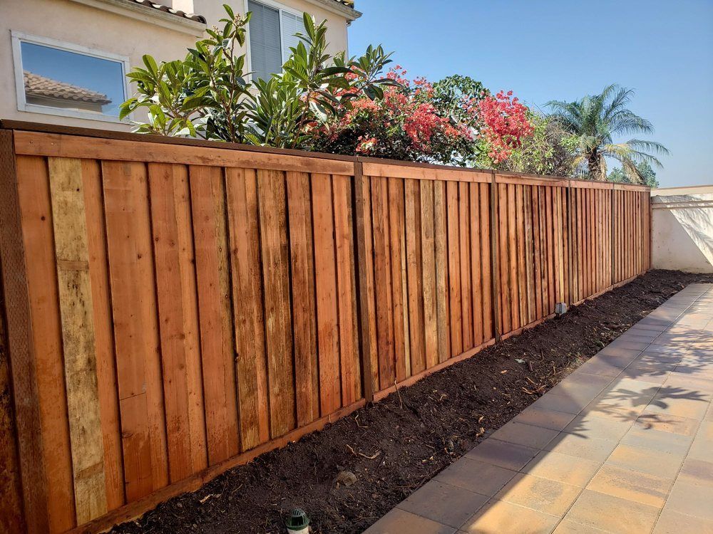 Wooden fence beside a tiled walkway, with garden shrubs and a house in the background