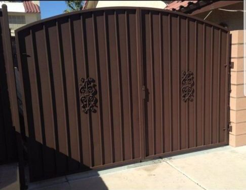 Brown metal driveway gate with decorative scrollwork panels in sunlight
