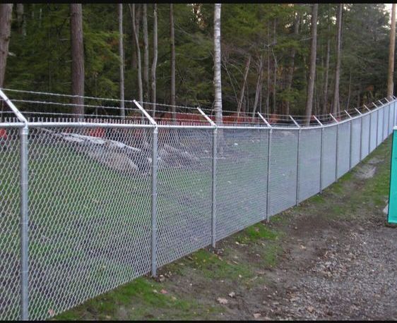 Chain-link fence along a gravel path beside a wooded area, with a blue gate at the far right.