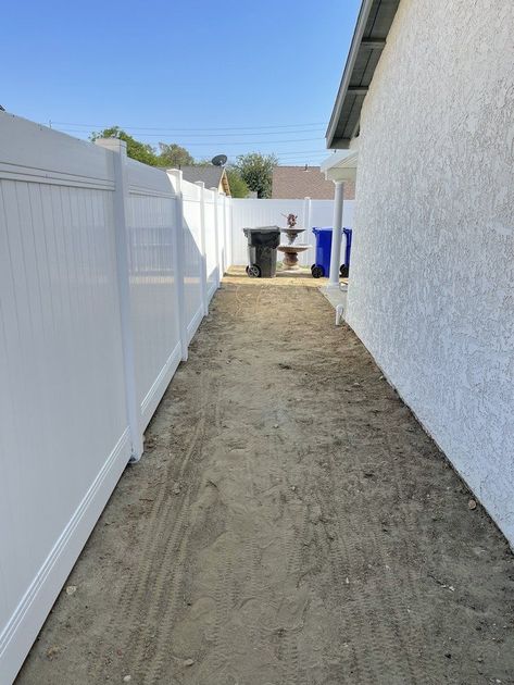 Narrow dirt side yard between a white fence and a gray house, with a small fountain at the end.
