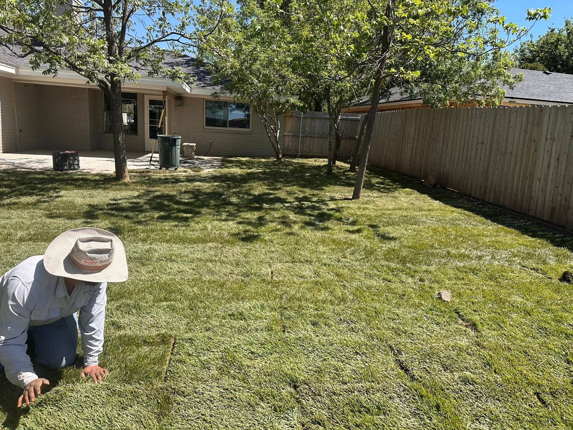 Person kneeling on a grassy backyard lawn beside a house and wooden fence, wearing a sun hat.