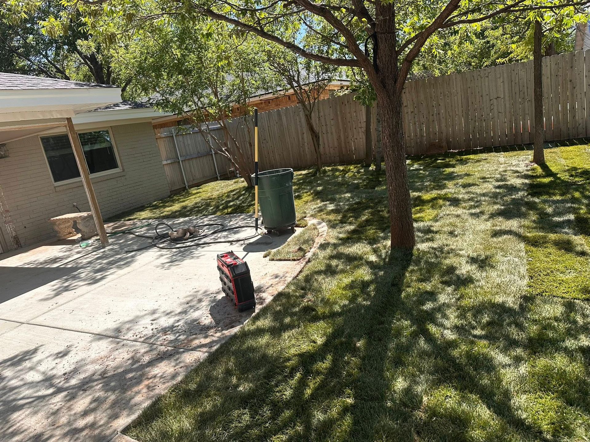 Backyard patio with a tree, wooden fence, house wall, and a red-and-black tool on the concrete.