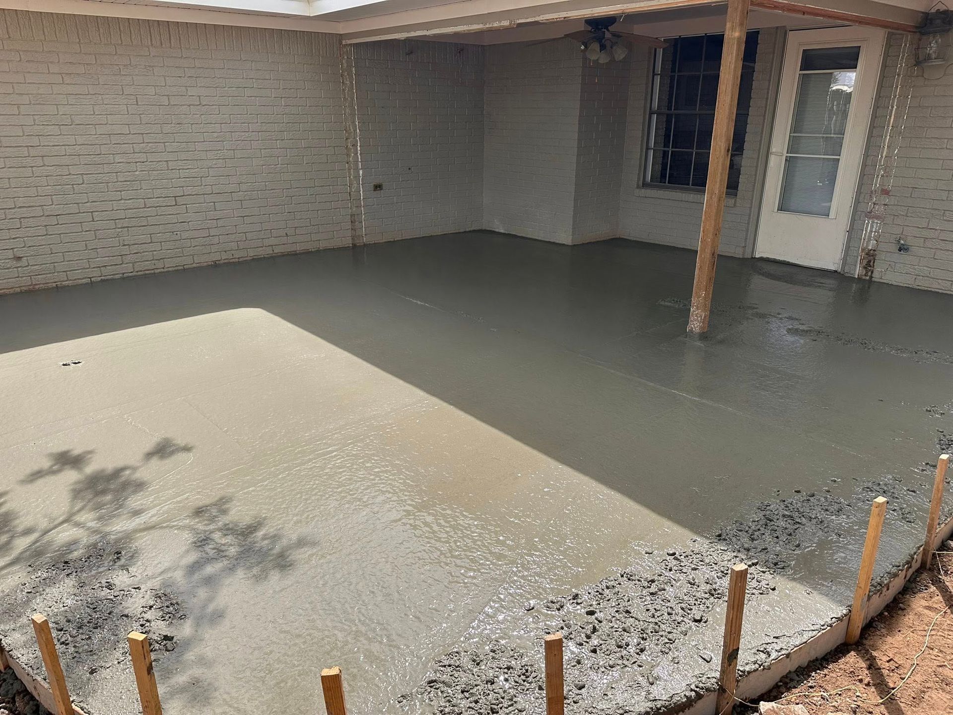 Freshly poured concrete slab under a covered patio beside a brick house, with wooden stakes around the edge