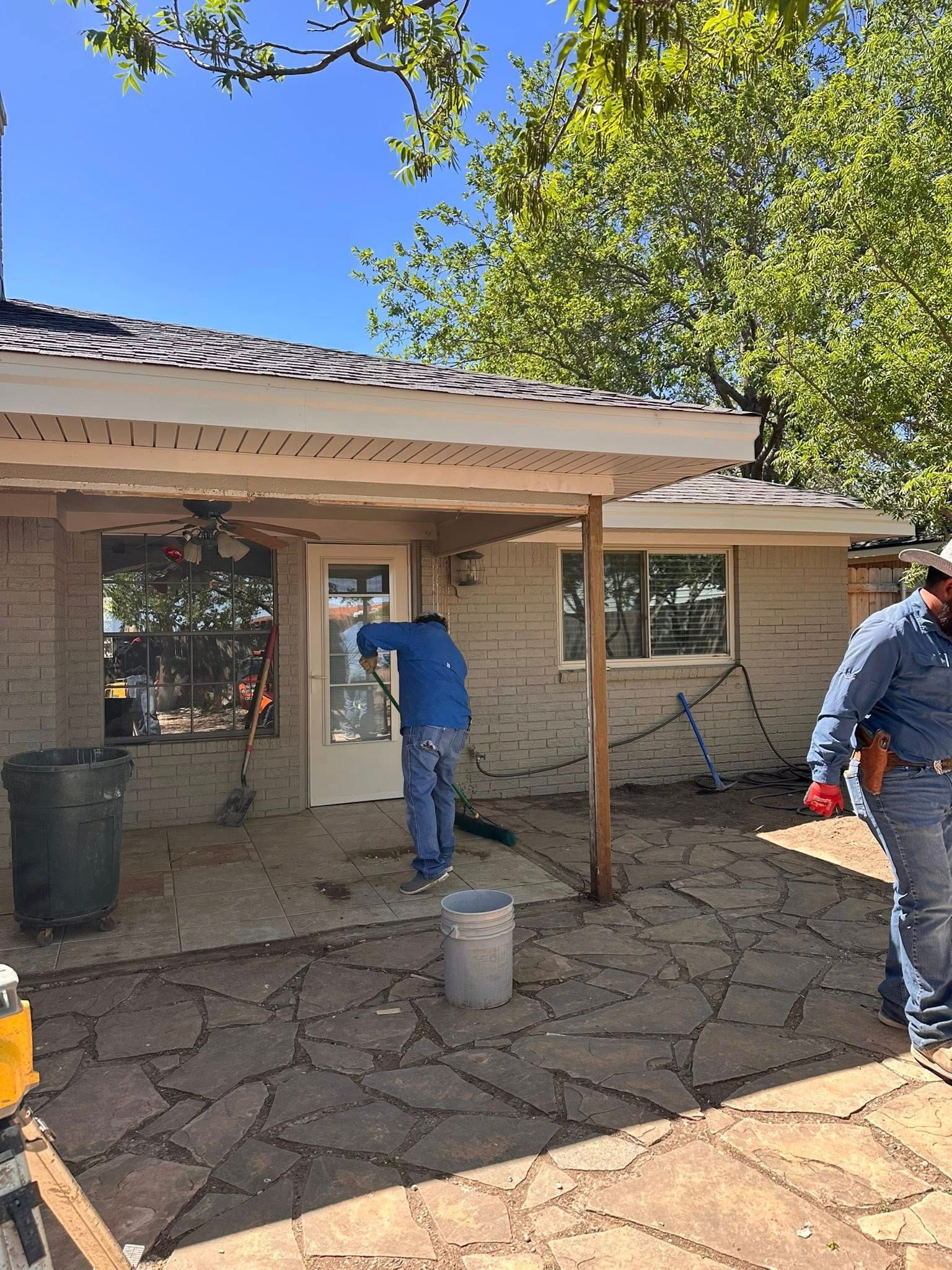 Workers painting an outdoor patio under a house overhang with a blue sky above