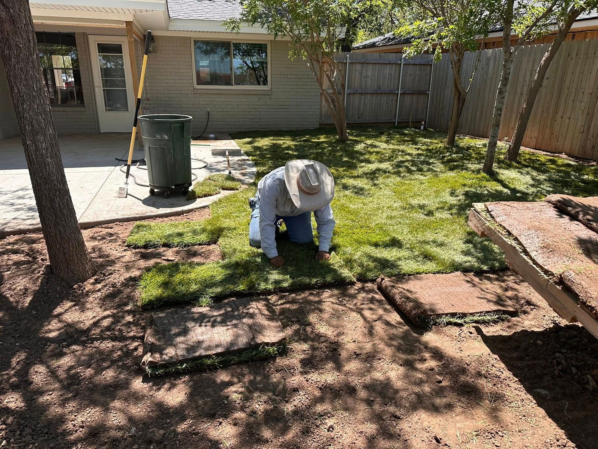 Person kneeling in a backyard beside a freshly laid green sod patch under bright sunlight