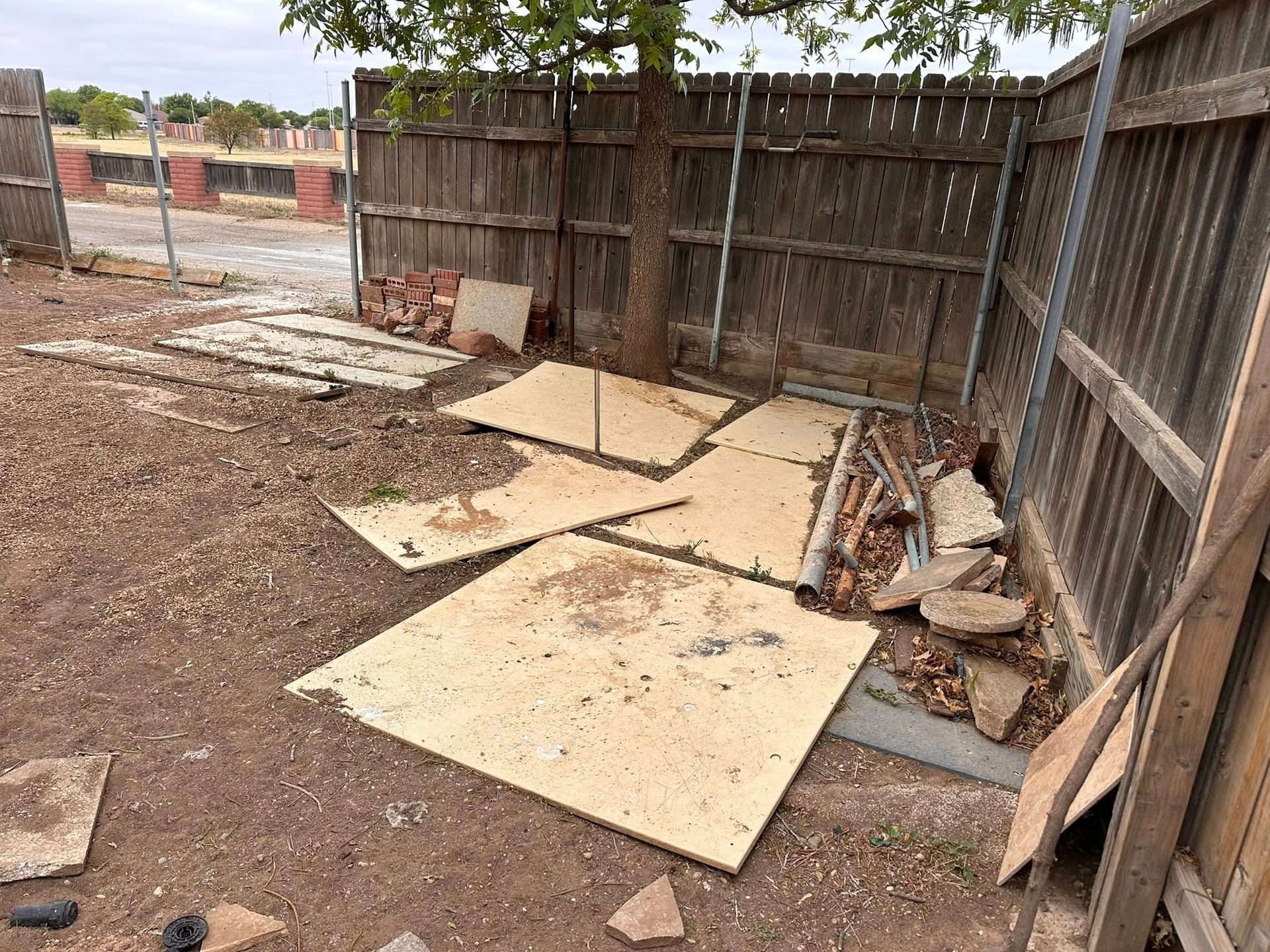 Dirt yard with scattered plywood boards and debris beside a weathered wooden fence