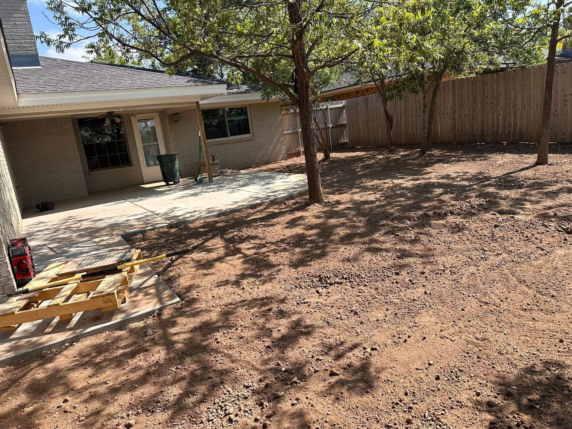 Backyard with a bare dirt yard, tree, wooden fence, and a house patio in the background