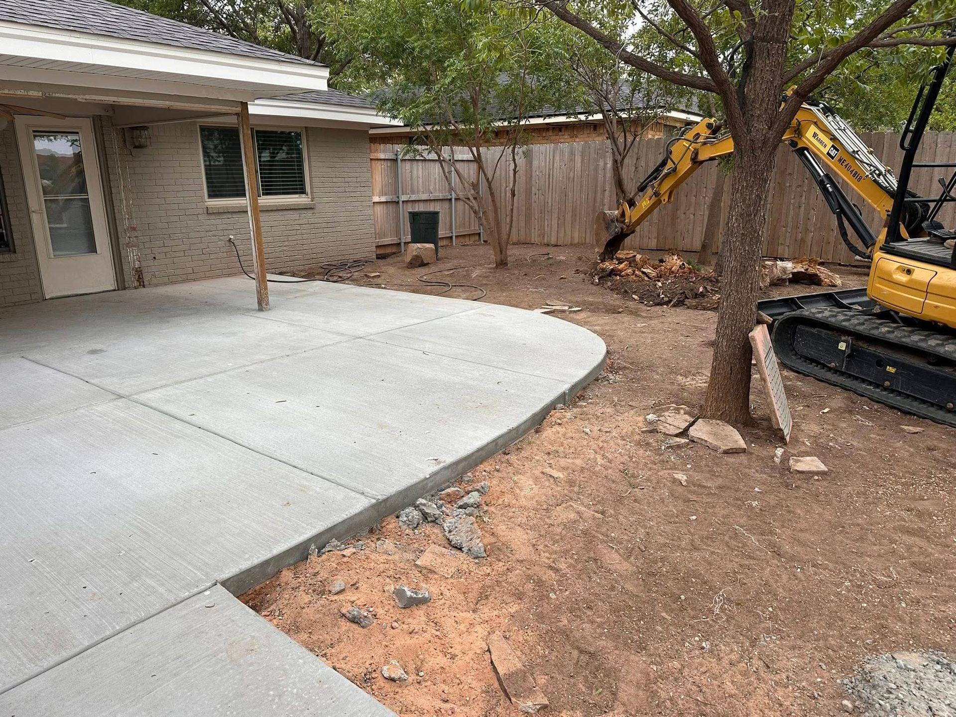 Backyard patio with fresh concrete slab beside a house, tree, and yellow excavator.