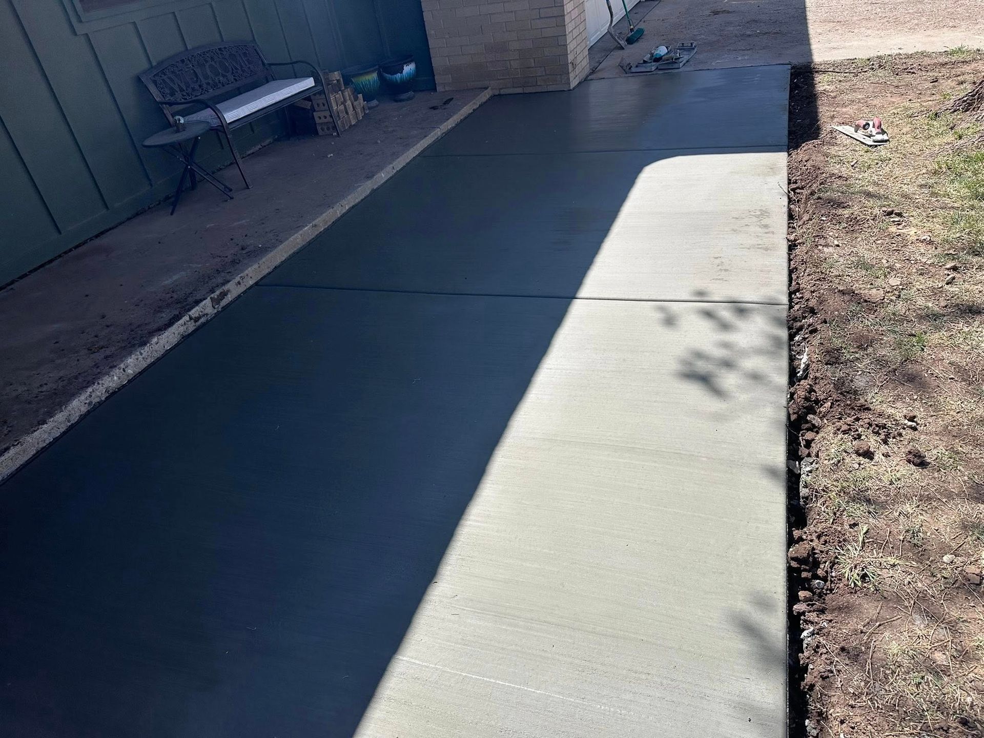 Freshly poured concrete walkway beside a house, partly shaded, with dirt and a bench nearby