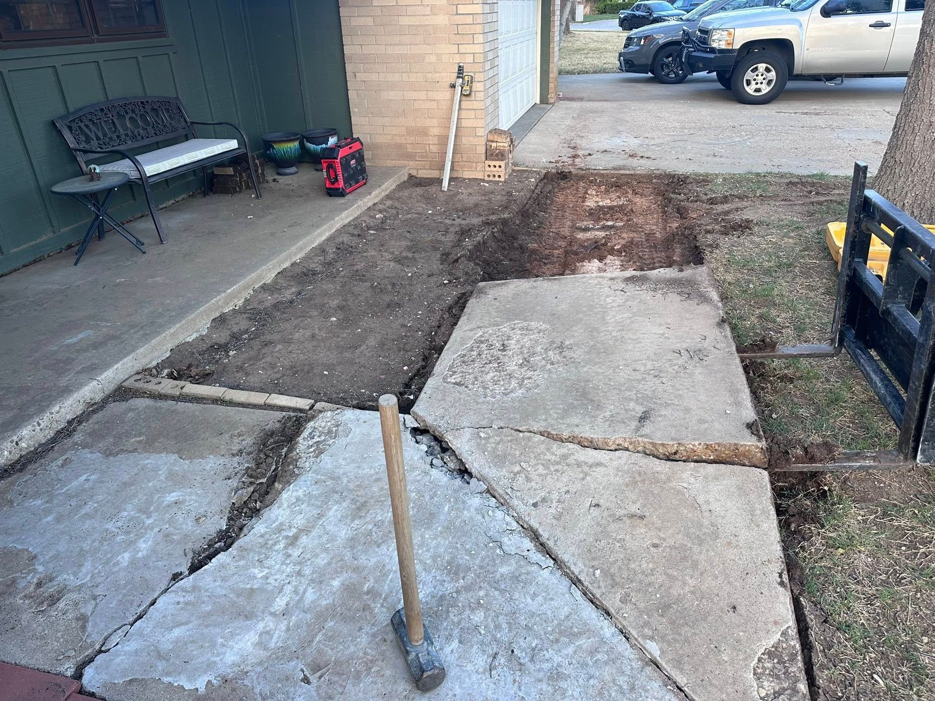 Damaged concrete walkway with broken slabs, dirt trenches, and tools beside a house driveway