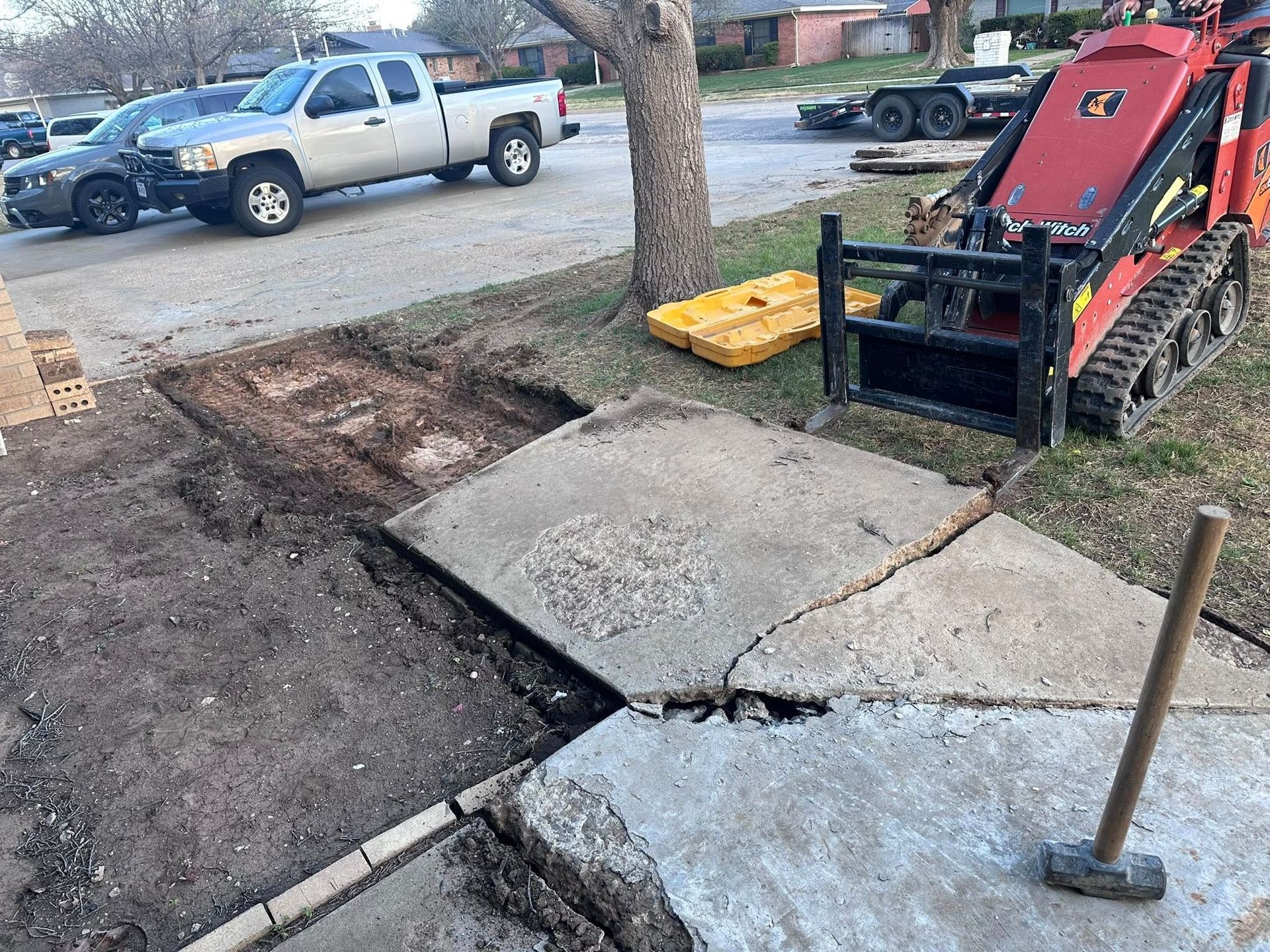 Damaged sidewalk with a broken concrete slab, dirt trench, and a tracked machine beside a pickup truck.