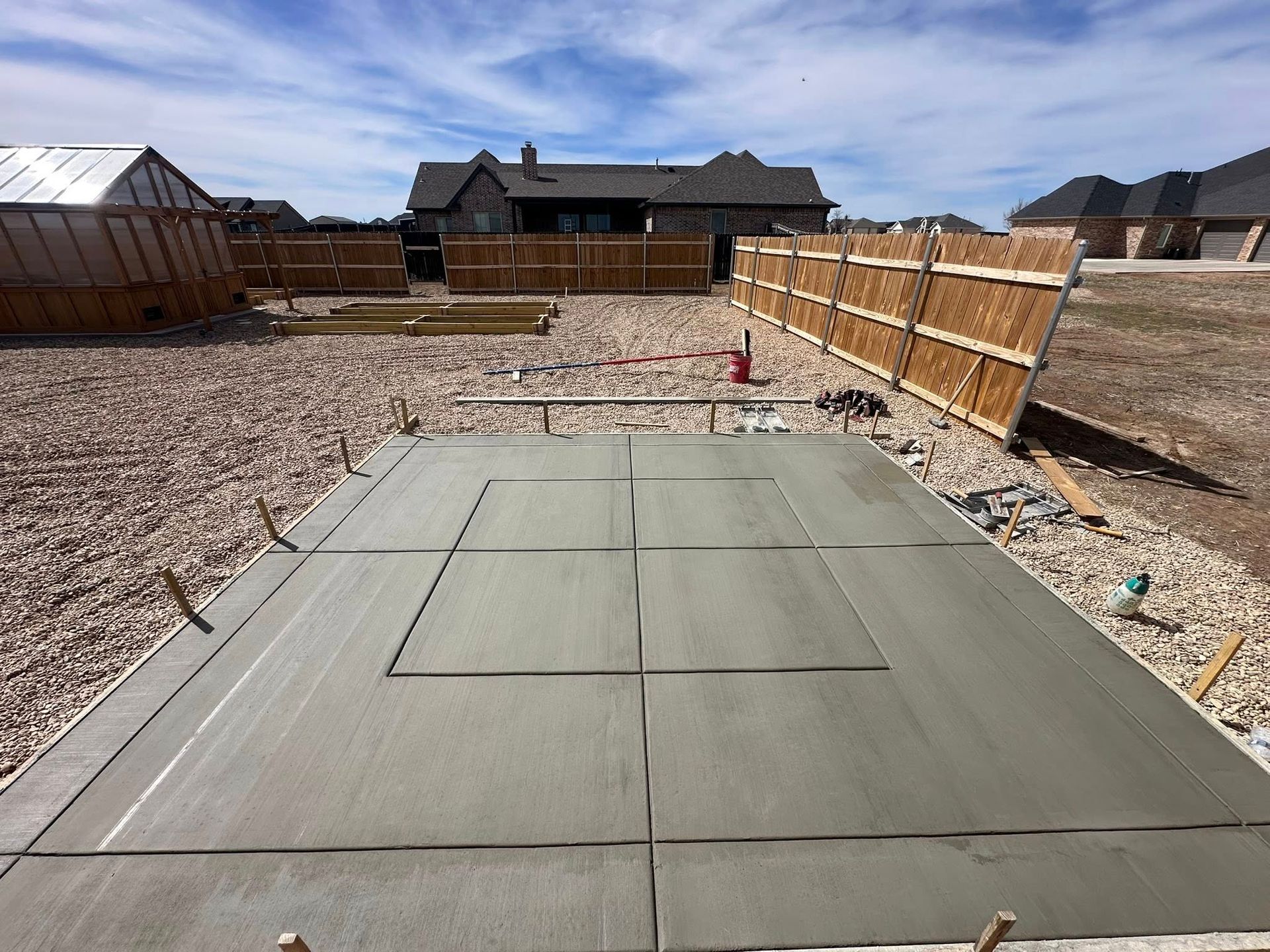 Concrete patio in a fenced backyard under construction, with a house in the distance.
