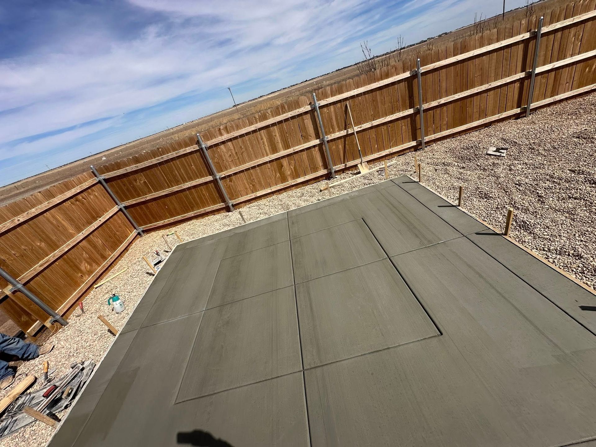 Concrete patio in a fenced backyard with gravel around the edges and a partly cloudy sky above