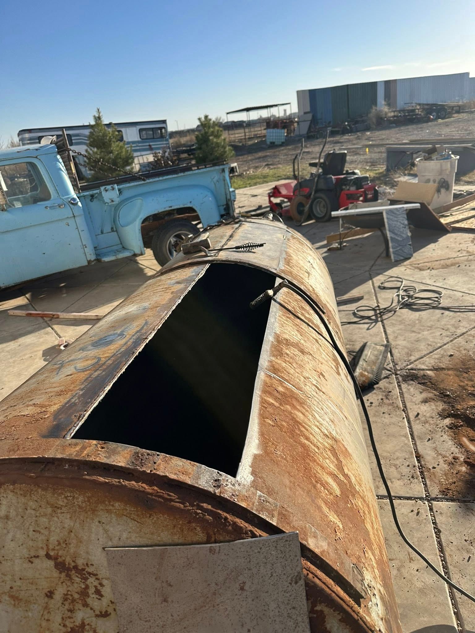 Rusty metal tank lying in an industrial yard, with vehicles and buildings in the background.