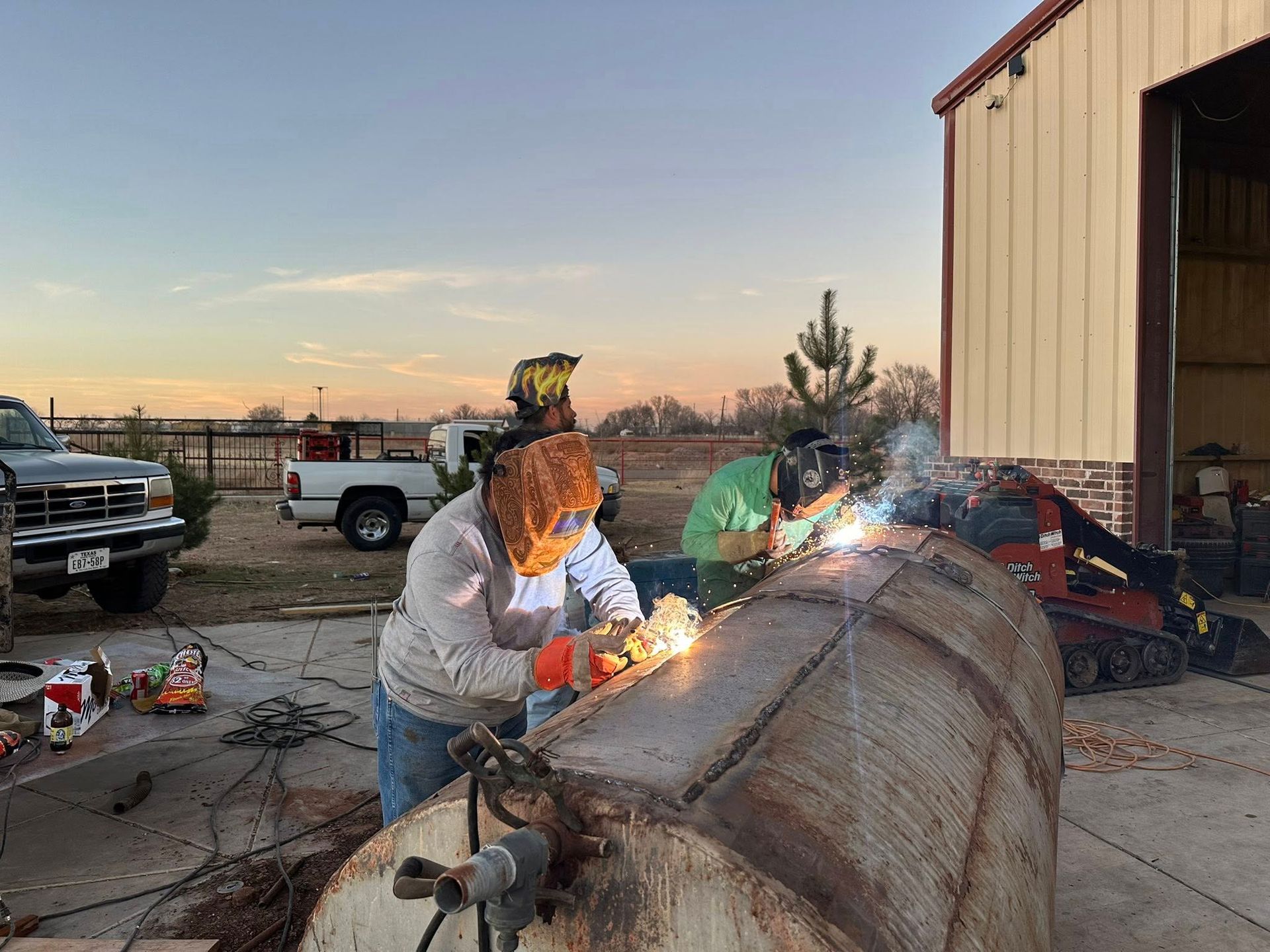 Two people welding a large metal tank outdoors beside a barn at sunset