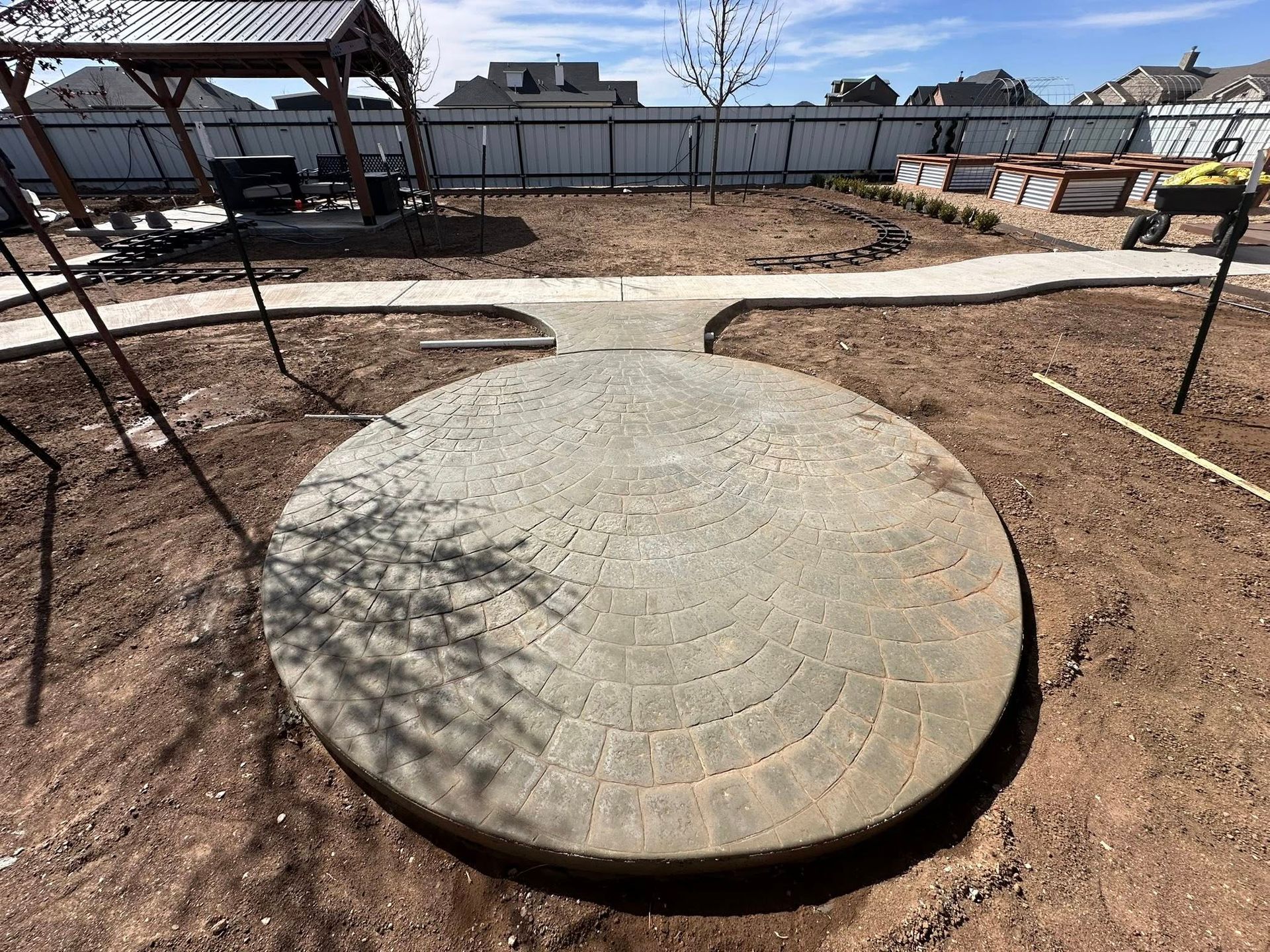 Concrete circular patio slab in a backyard, surrounded by dirt and a curved walkway near a fence.