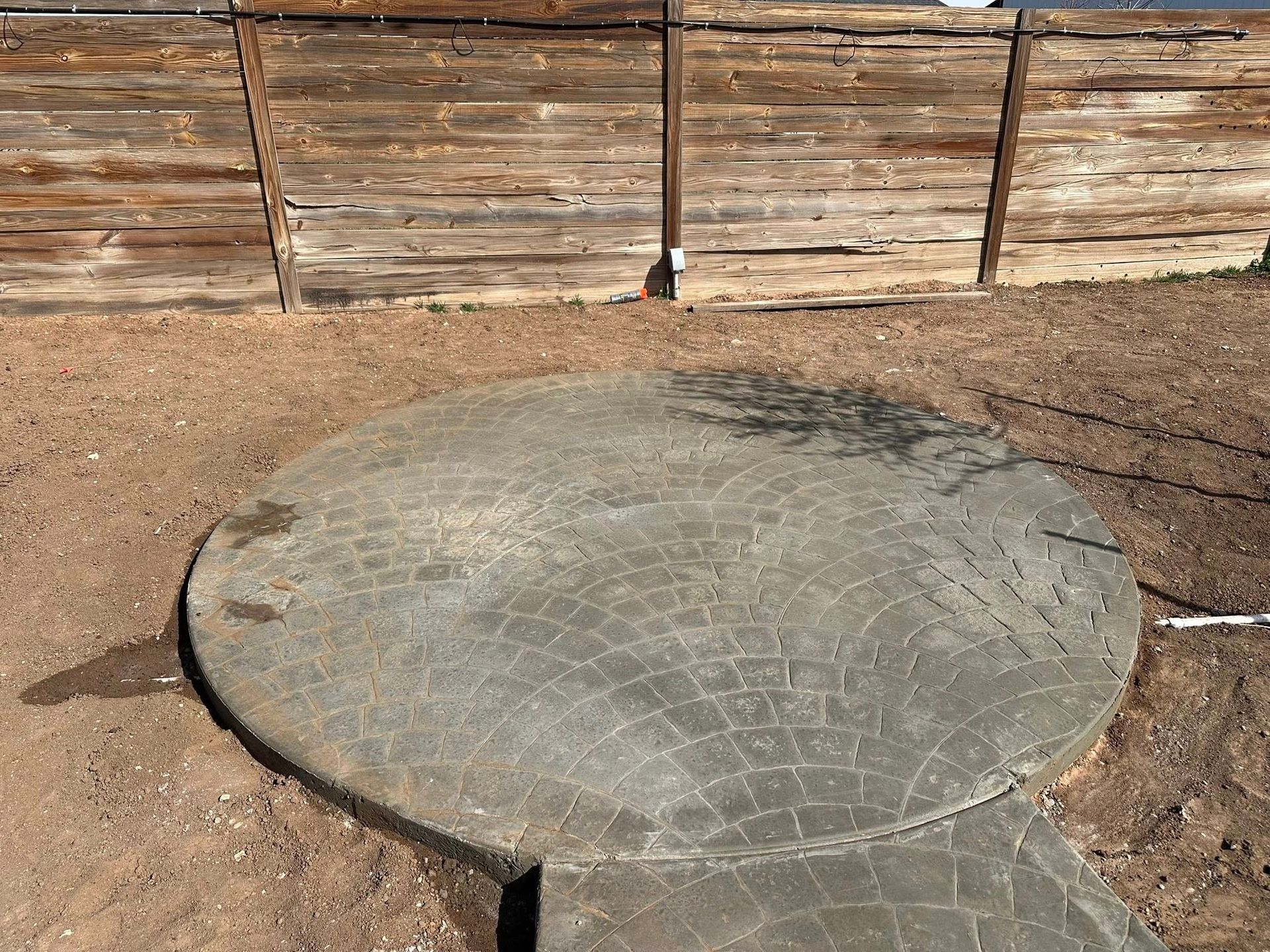 Concrete circular slab in a dirt yard in front of a weathered wooden fence.