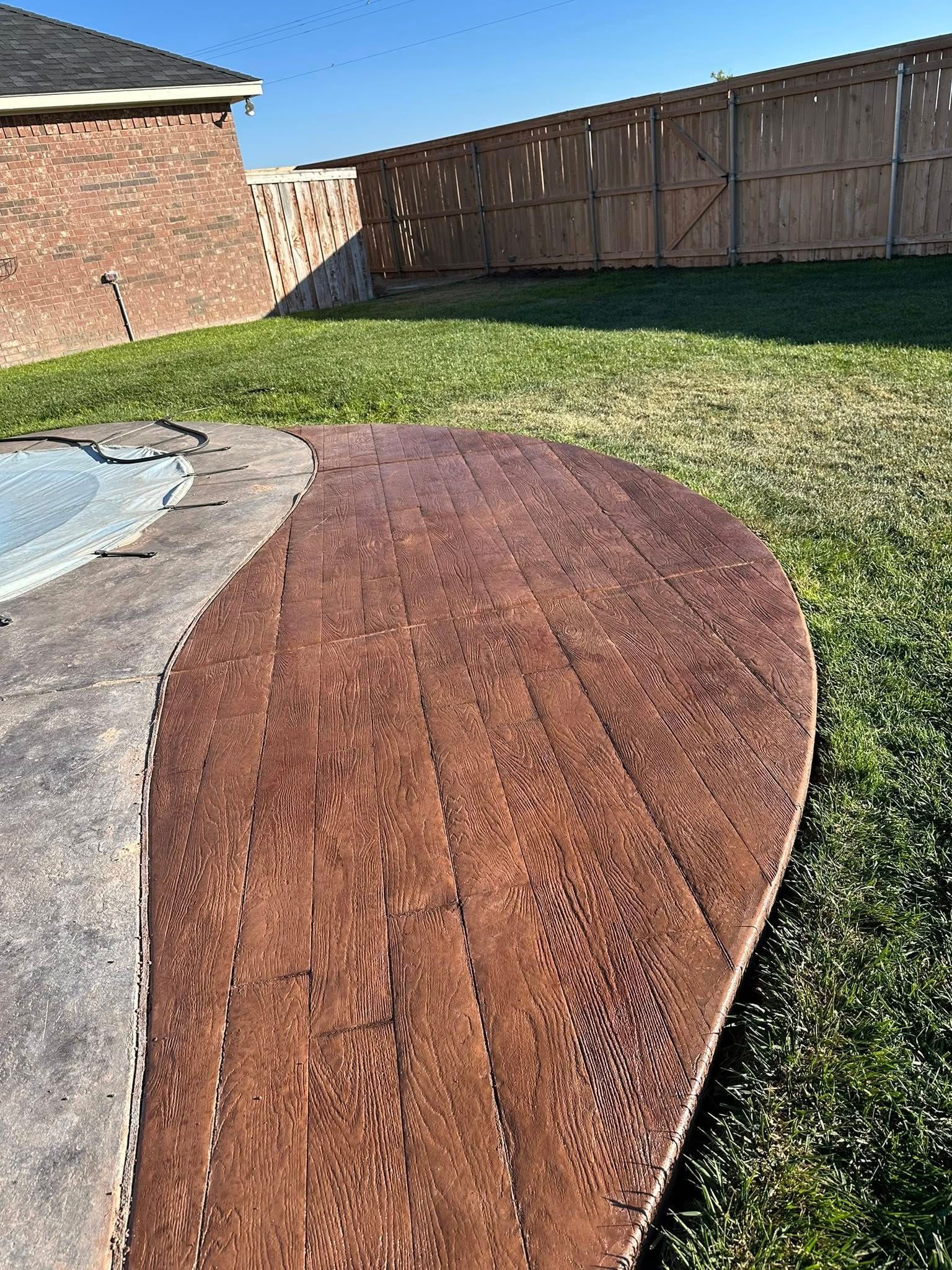 Rust-colored stamped concrete patio beside a grassy backyard and wooden fence.