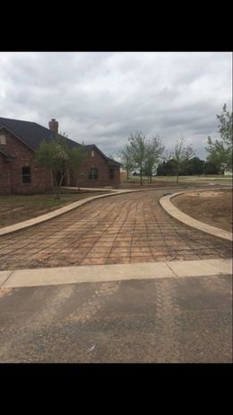 Curved brick-paved driveway beside a brick house under a cloudy sky, with trees and open land ahead